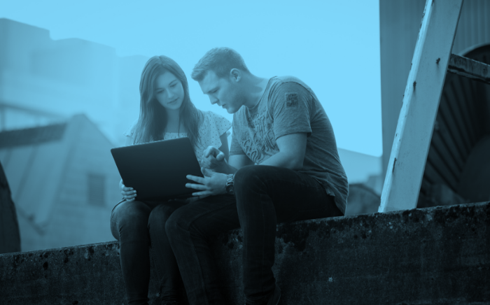 Studierenden sitzen auf einer Mauer und schauen in einen Laptop